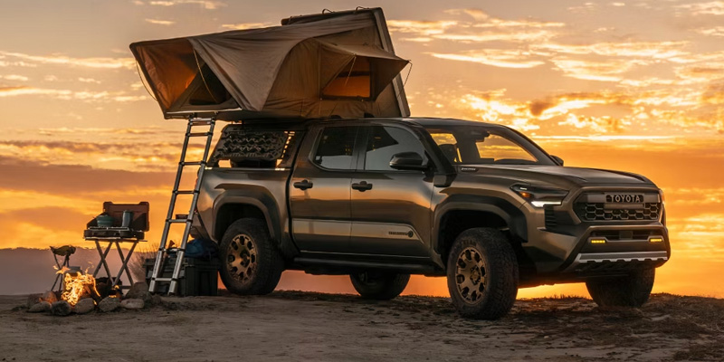 Toyota Tacoma aparcado en un camping con una tienda de campaña en el techo al atardecer, destacando su diseño robusto todoterreno y preparado para la aventura.