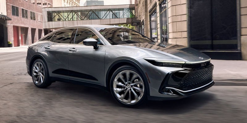 Vista frontal de un Toyota Crown plateado de 2025 circulando por una ciudad moderna