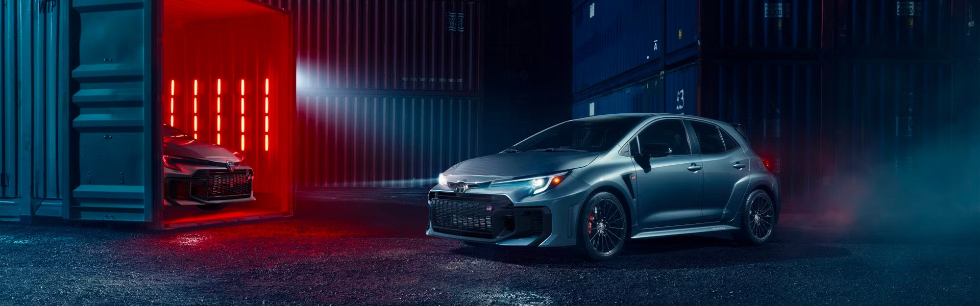 2025 Toyota GR Corolla en una dramática escena de almacén iluminado con un segundo vehículo dentro de un contenedor.