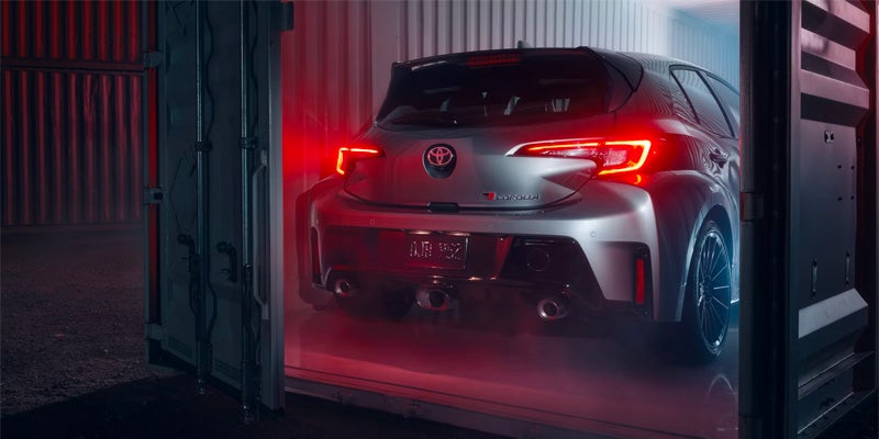 Vista trasera de un Toyota GR Corolla rojo de 2025 aparcado dentro de un contenedor de transporte con una iluminación espectacular.